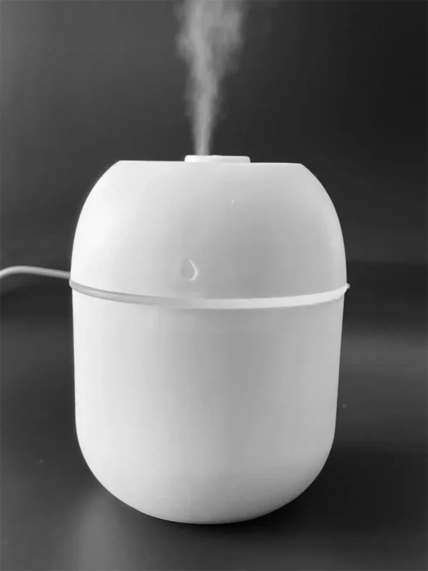 Forme: L'humidificateur a une forme arrondie, presque sphérique, ce qui lui donne un aspect compact et moderne. Couleur: Il est entièrement blanc, ce qui lui confère un aspect neutre et discret. Matériau: Le matériau semble être du plastique, ce qui le rend léger et facile à déplacer. Détails: On peut apercevoir un petit symbole représentant une goutte d'eau sur le dessus de l'appareil, ce qui indique clairement sa fonction. Il y a également un cordon d'alimentation visible, suggérant qu'il fonctionne avec une prise USB. Contexte: L'arrière-plan est noir uni, ce qui met en valeur la couleur blanche de l'humidificateur et permet de bien distinguer ses formes.