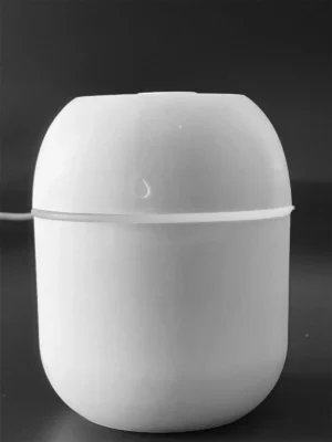 Forme: L'humidificateur a une forme arrondie, presque sphérique, ce qui lui donne un aspect compact et moderne. Couleur: Il est entièrement blanc, ce qui lui confère un aspect neutre et discret. Matériau: Le matériau semble être du plastique, ce qui le rend léger et facile à déplacer. Détails: On peut apercevoir un petit symbole représentant une goutte d'eau sur le dessus de l'appareil, ce qui indique clairement sa fonction. Il y a également un cordon d'alimentation visible, suggérant qu'il fonctionne avec une prise USB. Contexte: L'arrière-plan est noir uni, ce qui met en valeur la couleur blanche de l'humidificateur et permet de bien distinguer ses formes.